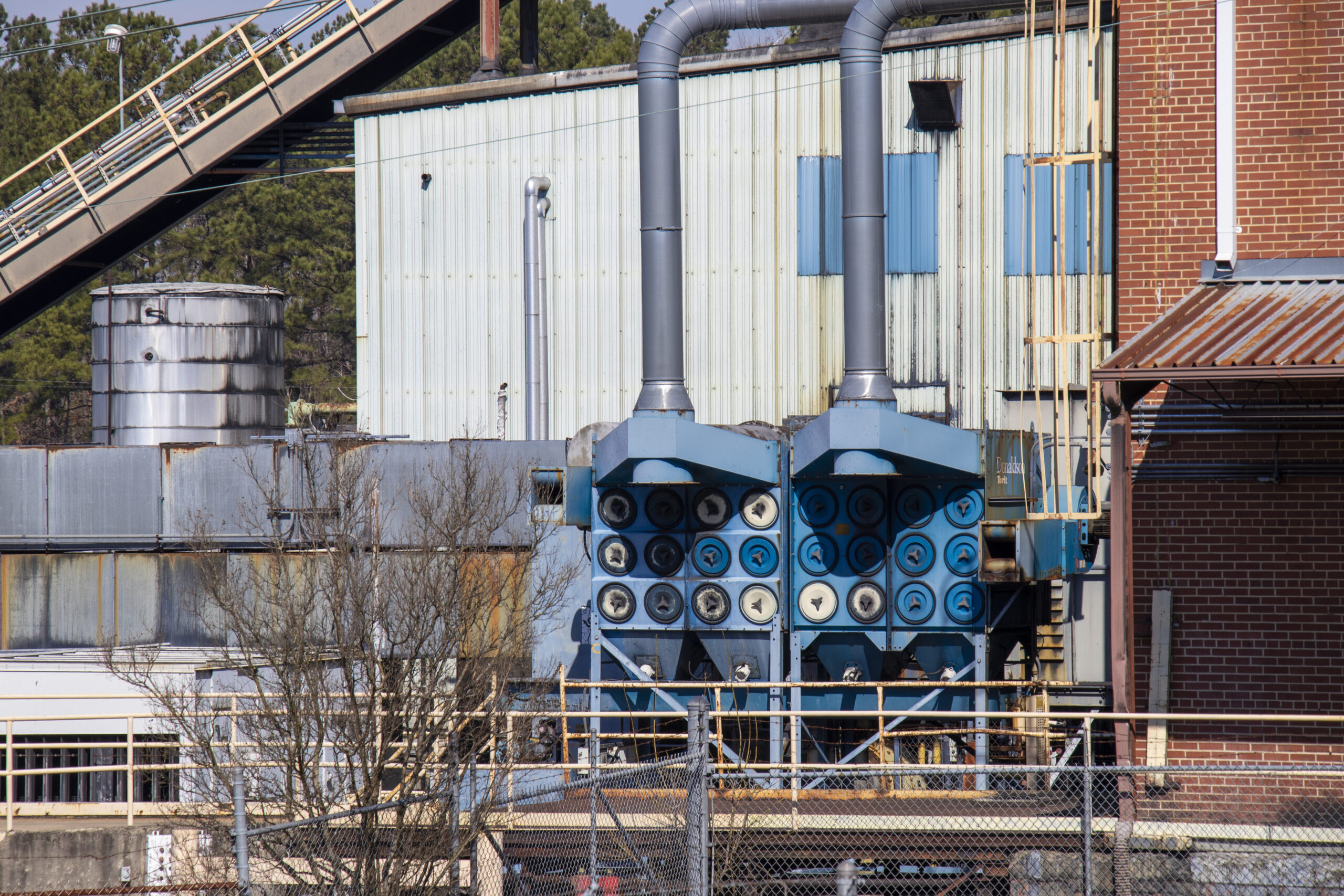 Exterior of a brick and metal industrial building with a dust collection system, conveyor, and storage tank behind a barbed wire fence.